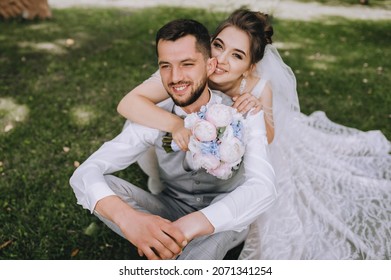 A beautiful, sweet bride in a dress hugs behind the back of a stylish, bearded, smiling groom in a gray vest and white shirt sitting on the green grass. Wedding portrait, photo of newlyweds in love.