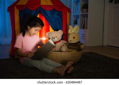 Beautiful Sweet Child Sitting In Front Of Kid Tent And Holding Comic Book Speaking Story For Her Favorite Toy In Bedroom At Night Before Going To Bed.