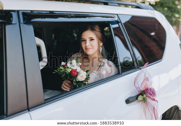 A beautiful, sweet curly-haired bride with\
a bouquet of flowers in a lace dress sits inside a white car.\
Wedding portrait of a brunette at the\
window.