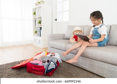 beautiful sweet kid girl sitting on living room sofa using mobile digital tablet pad searching travel information when she packing luggage suitcase at home.