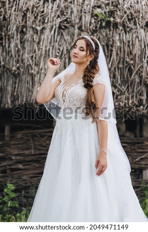 A beautiful, sweet, tender bride in a white dress with a veil and a diadem on her head, standing in the village against the background of a hut with a straw. Wedding photography close-up, portrait.