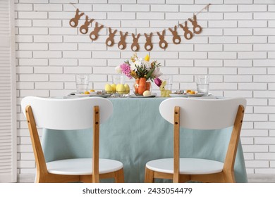 Beautiful table setting for Easter decorated with bouquet of carrots and flowers near white brick wall