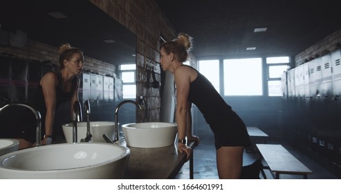 Beautiful Tall Sporty Blonde Woman Washing Face, Looking In The Mirror In Dark Gym Locker Room Exhausted Slow Motion.