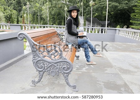 Beautiful tan skin Asian chic woman work with phone and hand write note on dairy book with hot coffee cup to go .Presenting your product. Businesswoman sit on wooden bench in flower garden.