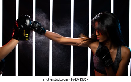 Beautiful Tanned skin Asian Woman is training and punching with Black Mitts Gloves with Coach pads. Office Girl exercises in Modern Neon Muay Thai Boxing Gym heavily with sweat water splash in air