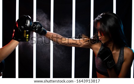 Beautiful Tanned skin Asian Woman is training and punching with Black Mitts Gloves with Coach pads. Office Girl exercises in Modern Neon Muay Thai Boxing Gym heavily with sweat water splash in air