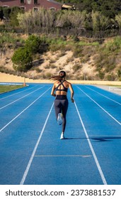Beautiful tanned slim young runner girl, dressed in tight sportswear, running and sprinting on her back with her tail waving in the wind on a blue athletics track.