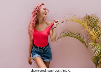Beautiful tanned woman playfully posing beside palm tree and laughing. Indoor portrait of fascinating girl with pink hair wears denim shorts and tank-top.