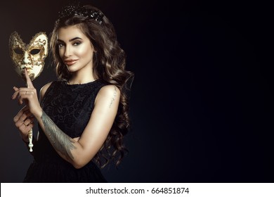 Beautiful tattooed smiling lady with dark wavy shining hair and perfect make-up wearing lace dress and jewel black crown holding a Venetian mask in her hand with silence gesture. Copy space
