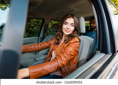 Beautiful Taxi Driver Sitting In A Car And Smiling At The Camera With Passenger In The Background. 