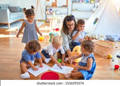 Beautiful Teacher And Group Of Toddlers Sitting On The Floor Drawing Using Paper And Pencil Around Lots Of Toys At Kindergarten