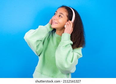 beautiful teen girl wearing green sweater standing against blue background wears stereo headphones listens music concentrated aside. People hobby lifestyle concept