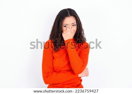 beautiful teen girl wearing knitted red sweater over white background , holding his nose because of a bad smell.