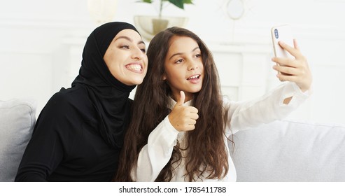 Beautiful Teen Small Girl And Young Arabian Mother In Hijab Sitting On Couch And Taking Selfie Photos With Smartphone Camera At Home. Arab Woman With Daughter Making Pictures Selfies With Mobile Phone