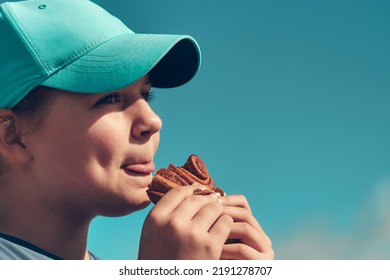 Beautiful teenage girl eating a cinnamon bun, side view. Child snacking outdoors, face close-up