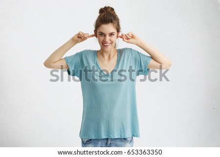 Beautiful teenage girl wearing jeans and oversize blue t-shirt plugging ears with fingers and smiling broadly, teasing, pretending not to lsiten, ignoring someone. Human emotions and feelings