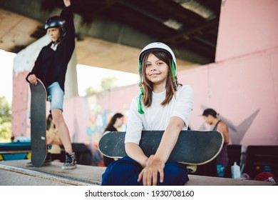 Beautiful teenage girl in white helmet sits with skateboard in her hands on pipe ramp in an extreme sports park in city during training in sports club. Young female skateboarder on half pipe ramp. - Powered by Shutterstock