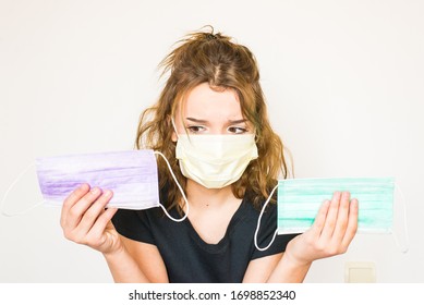 Beautiful Teenager Brown-eyed Girl With Brown Hair In Black T-shirt,wearing Mask,showing Two Coloured Hygienic Masks In Hand,looking At Masks With Worried Eyes.Mask Collection. Mask Variety. Copyspace