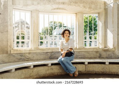 Beautiful Teenager Girl, With Brown Curly Hair, In Jeans And Blouse, With Small Bag, Sits On Bench In Park Gazebo, In Baroque Style, Waiting For Meeting, In Sun