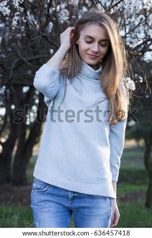 Beautiful tender girl touches her hair. Pretty young woman stands in park on blossom trees background. She dressed in white pullover and blue jeans she looking down and smile. Medium long shot.