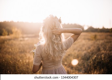 Beautiful Tender Happy Bride With Blonde Curly Hair And A Head Wreath Standing In A Sunset Summer Field, Enjoying Herself, Standing Background To The Camera