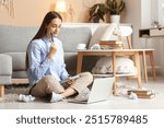 Beautiful thoughtful female writer with laptop and notebook sitting on floor at home