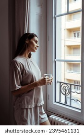 Beautiful thoughtful girl with long hair standing in bedroom looking at window drinking cofffee. Young woman feeling lonely looking at neighbor windows  - Shutterstock ID 2395461131