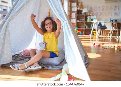 Beautiful Toddler Wearing Glasses And Unicorn Diadem Sitting On The Floor At Kindergarten Celebrating Mad And Crazy For Success With Arms Raised And Closed Eyes Screaming Excited. Winner Concept