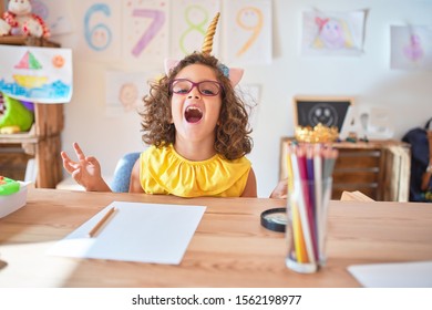 Beautiful Toddler Wearing Glasses And Unicorn Diadem Sitting On Desk At Kindergarten Crazy And Mad Shouting And Yelling With Aggressive Expression And Arms Raised. Frustration Concept.