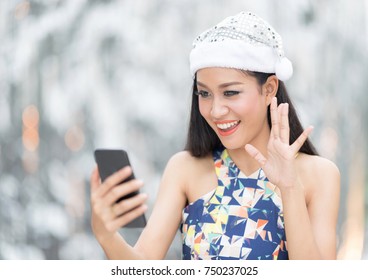 Beautiful Toothy Smiling Young Woman In White Santa's Hat  Using Smartphone Doing Facetime With Family Against Blur Background.Christmas Concept.