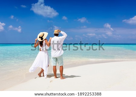 A beautiful tourist couple in white summer clothes and hats stands on a tropical beach and gazes at the turquoise ocean during their vacation time