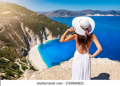 A beautiful tourist woman in white dress and sunhat enjoys the view to the famous Myrtos Beach on the island of Kefalonia, Ionian Sea, Greece