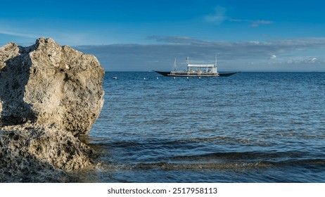 The beautiful traditional Filipino bangka boat is anchored in the turquoise ocean. Lifebuoys are on board. Masts against the blue sky and clouds. A picturesque boulder in the foreground. Philippines.  - Powered by Shutterstock