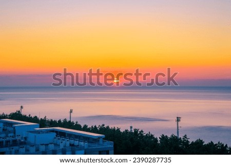 Beautiful and tranquil sunrise seascape over Sokcho Beach in Sokcho City, Gangwon Do Province, South Korea, with orange colored clouds, horizon, pine tree forest, rooftop view and beach light pylons