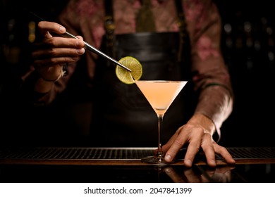 beautiful transparent martini glass with bright cocktail on the bar counter and hand of bartender holds slice of lemon with tweezers and decorates the glass with it