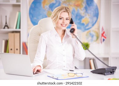 Beautiful Travel Agent Sitting At The Table In Office And Talking On Phone.