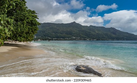 Beautiful Tropical Beach. The Waves Of The Turquoise Ocean Are Foaming On The Sand. Lush Vegetation On The Shore. A Picturesque Boulder In The Water. A Green Hill Against A Blue Sky And Clouds.