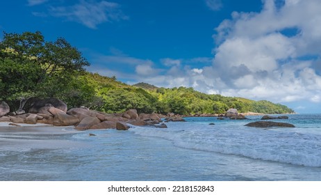 Beautiful Tropical Beach. The Waves Of The Turquoise Ocean Spread And Foam On The Sand. Picturesque Boulders In The Distance. A Green Hill On A Background Of Blue Sky And Clouds. Seychelles. Praslin.