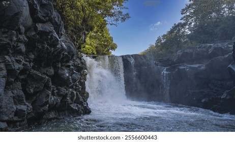 A beautiful tropical waterfall. Streams of water flow from the edge of the ledge into the river. Foam, splashes. There is green vegetation on the steep rocky shores. Blue sky, clouds. Mauritius. - Powered by Shutterstock