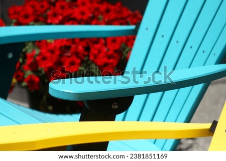 Beautiful turquoise bright blue Muskoka Adirondack chair. Arm of yellow chair. Chairs. Comfort. Relaxation. Leisure. Comfortable. Barrel of red petunia flowers. Summer. Outdoors. Concept. Cottage.