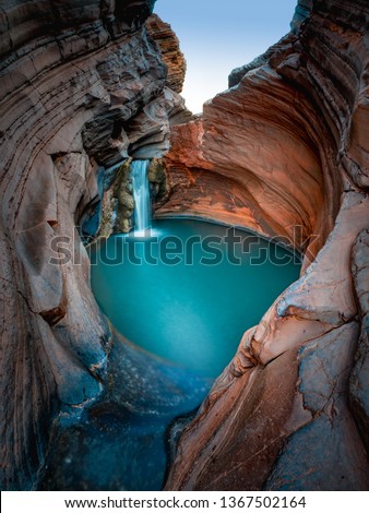 Beautiful turquoise waterfall flows into natural pool surrounded by red rock formation in Karijini National Park in Western Australia.