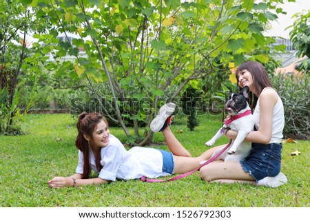 beautiful two young asian woman wearing white shirt who lying on grass and playing with her cute dog with happy and smiling face in garden with green trees. (friendship concept)