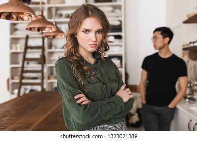 Beautiful Upset Woman With Wavy Hair Holding Hands Together Angrily Looking In Camera With Asian Man On Background. Young International Couple Quarreling Spending Time On Kitchen At Home