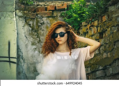 Beautiful Vape Teenager. Portrait Of A Pretty Young White Girl With Red Curly Hair In Sunglasses Vaping An Electronic Cigarette Near The Old Ruined Village House. Lifestyle.