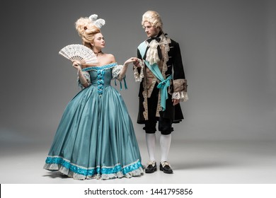 beautiful victorian woman with fan looking at gentleman in wig while holding hands on grey