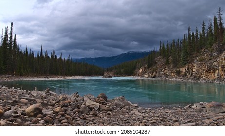 Beautiful view of Athabasca River in a valley in the Rocky Mountains surrounded by coniferous forest with stones in front in Jasper National Park, Alberta, Canada on cloudy day in autumn season.