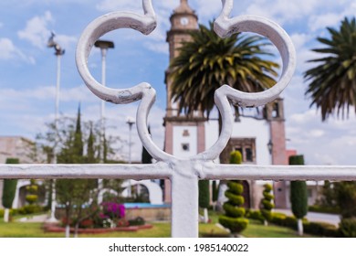 Beautiful View From Behind White Metal Fence Of Old Hispanic Church Surrounded By Bushes And Palm Trees. Ancient Bell Tower With Slightly Cloudy Sky As Background. Classic Mexican Architecture