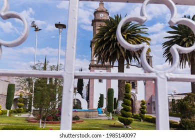 Beautiful View From Behind White Metal Fence Of Old Hispanic Church Surrounded By Bushes And Palm Trees. Ancient Bell Tower With Slightly Cloudy Sky As Background. Classic Mexican Architecture