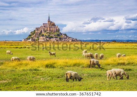 Beautiful view of famous historic Le Mont Saint-Michel tidal island with sheep grazing on fields of fresh green grass on a sunny day with blue sky and clouds in summer, Normandy, northern France