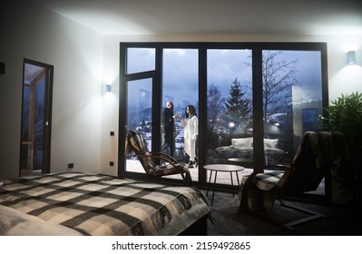 Beautiful View Of Man And Woman Toasting With Wine Behind Panoramic Window Of Scandinavian House Barnhouse. Interior Of Wooden House Room With Chairs, Bed And Couple In Love Outside Building.